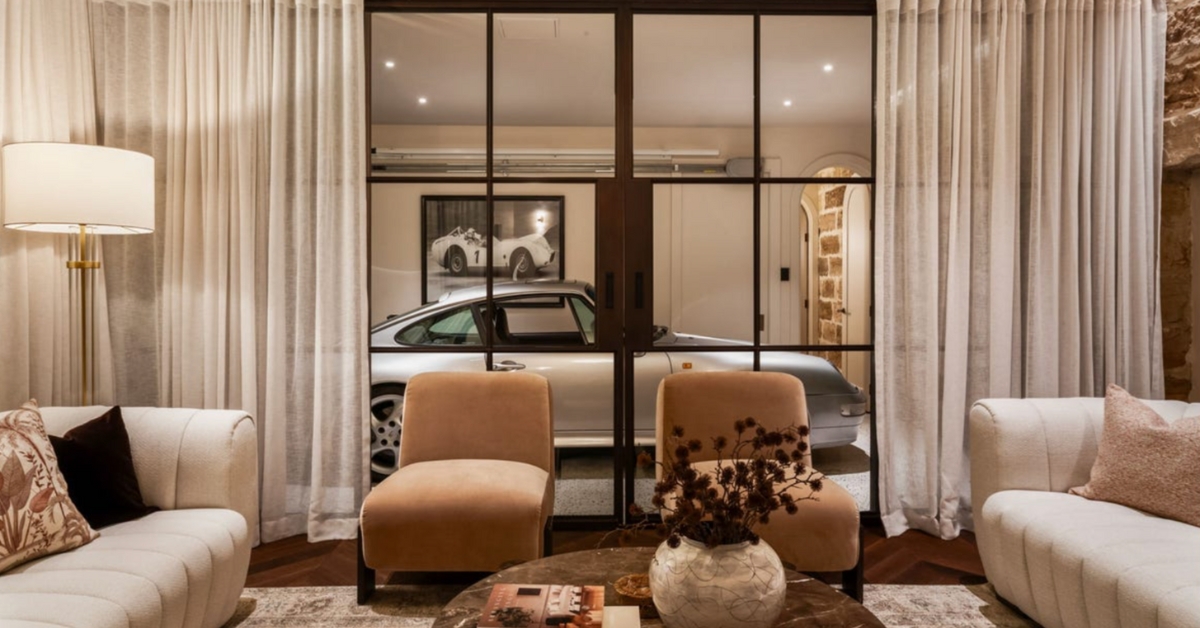 A sophisticated living room design featuring high-end renovation styling furniture with white textured sofas and tan armchairs, set on herringbone floors and overlooking a glass-enclosed garage housing a silver Porsche.
