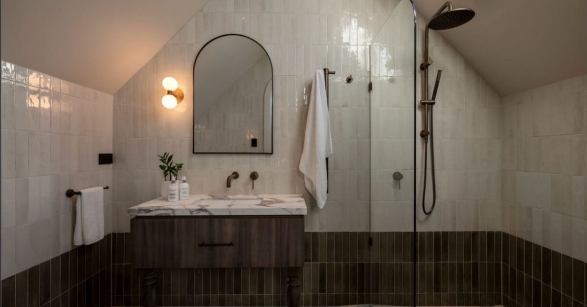 Completed bathroom renovation featuring the 12V wall sconce light fixture preserved by the 12V conversion hack, positioned next to an arched mirror with white and dark vertical tiles and antique brass shower fittings under a sloped ceiling.