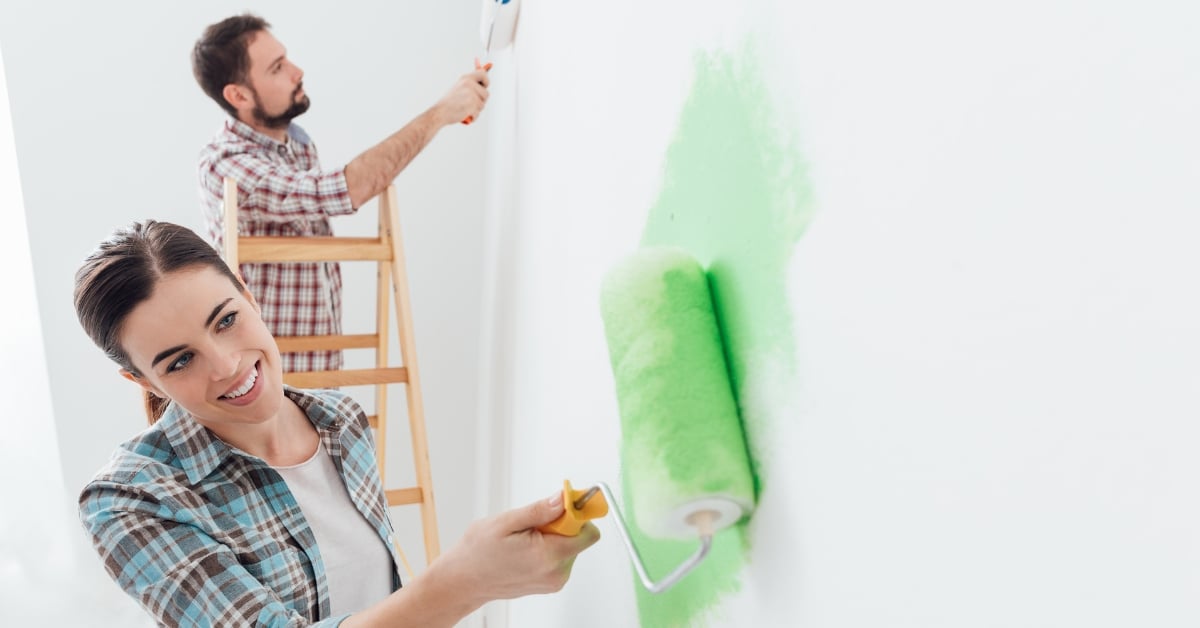 A smiling couple painting a wall green, illustrating how using renovation skills strategically can help fix fundamental problems in life like financial insecurity.