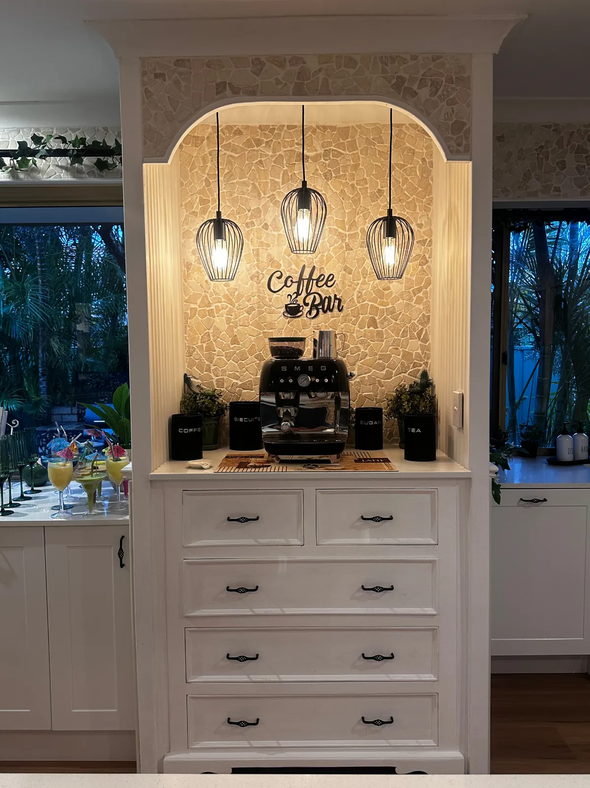 Arched coffee bar with a stone feature wall, three cage pendant lights, an espresso machine and white drawers
